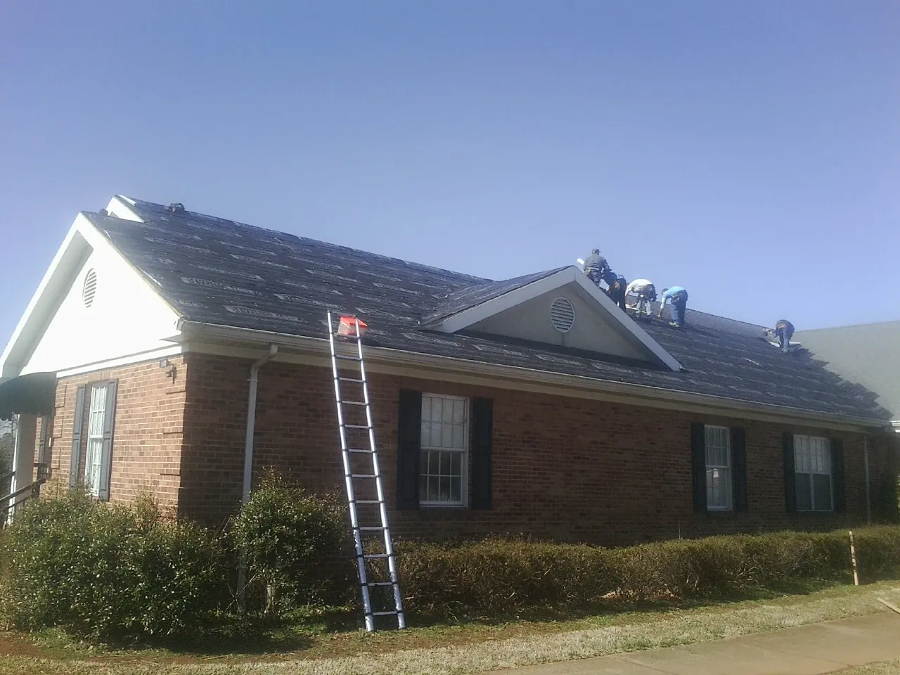 Crew tearing shingles off a brick ranch roof with a ladder set — Metal Roof Repair work by our crew in Lando