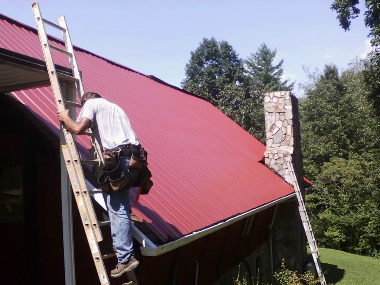 Roofer on a ladder beside a red metal roof and stone chimney — Soffit & Fascia Repair by Roofing Brothers in Lando